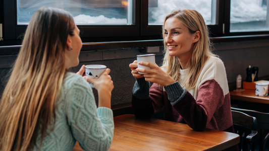 A person  smiles while holding a cup of coffee, engaged in conversation with friends at a cafe