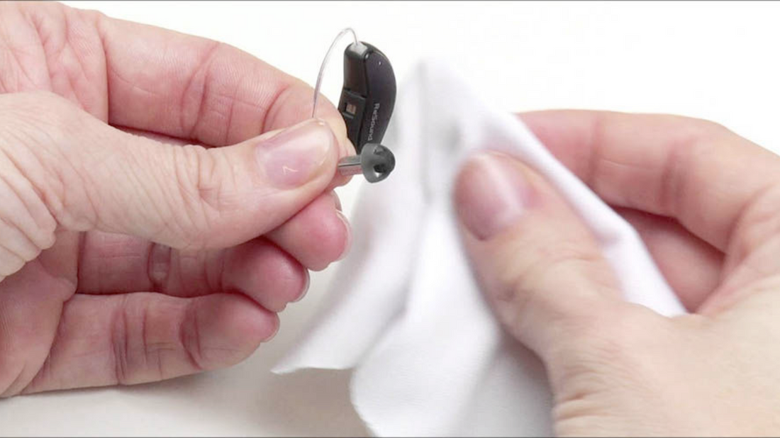 Illustration showing a person gently cleaning a behind-the-ear hearing aid with a small brush, with other cleaning tools and accessories in the background.